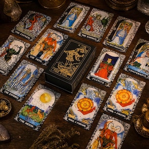 May include: A black tarot card deck box with gold foil illustration of The Fool, surrounded by various tarot cards including The Empress, The Devil, The High Priestess, Justice, Temperance, The Moon, and Wheel of Fortune on a dark surface.