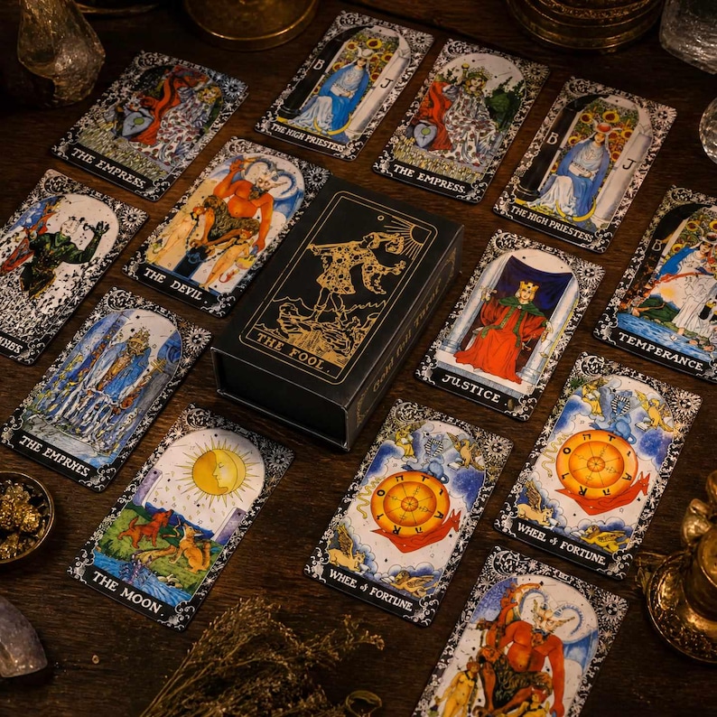 May include: A black tarot card deck box with gold foil illustration of The Fool, surrounded by various tarot cards including The Empress, The Devil, The High Priestess, Justice, Temperance, The Moon, and Wheel of Fortune on a dark surface.
