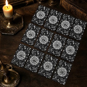 May include: A set of eleven rectangular tarot cards arranged in a grid, each featuring a black background with a white celestial sun illustration surrounded by stars and constellations. A lit candle in a brass holder sits to the left of the cards.