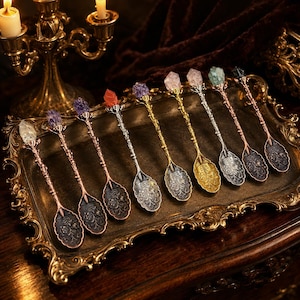 Puede incluir: Una colección de ocho cucharas de metal dispuestas en fila, cada una con un cuenco adornado con relieves florales, un tallo texturizado y una punta de cristal en bruto en el mango. Las cucharas varían en acabados metálicos como oro, plata y cobre.