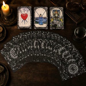 May include: A deck of tarot cards with black backs featuring white celestial star patterns, fanned out alongside three upright cards: Three of Swords, Two of Swords, and The Chariot. A lit candle and a crystal ball are visible in the dark setting.