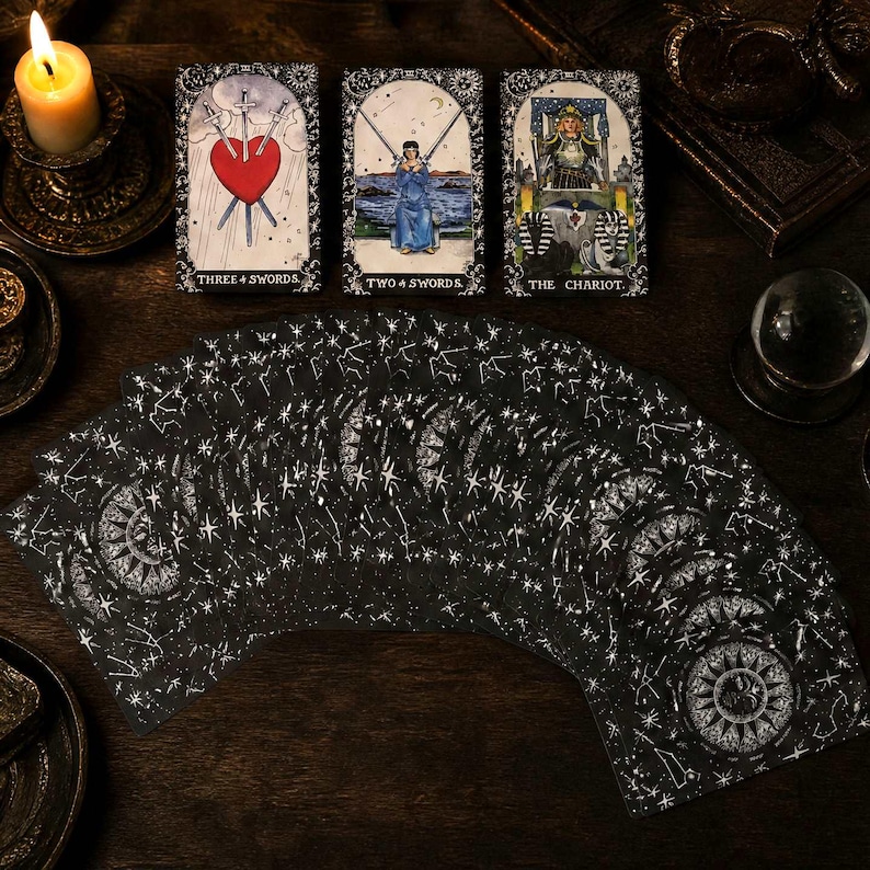 May include: A deck of tarot cards with black backs featuring white celestial star patterns, fanned out alongside three upright cards: Three of Swords, Two of Swords, and The Chariot. A lit candle and a crystal ball are visible in the dark setting.