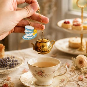 Puede incluir: Un infusor de té de metal dorado con forma de tetera en miniatura, suspendido por una cadena de un dije decorativo que muestra dos tazas de té azul claro y blanco sobre un platillo, posicionado sobre una taza de porcelana con flores.