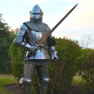 May include: A full suit of polished steel plate armor featuring a rounded helmet with a visor, a chainmail aventail, articulated arm and leg defenses, and metal gauntlets. The armor is holding a long, straight steel sword with a cross-shaped hilt.