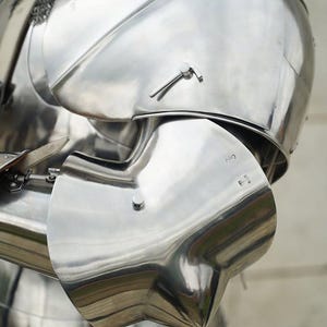 May include: A polished steel suit of armour featuring a rounded shoulder pauldron and a curved elbow couter. The metallic surface reflects light, showing smooth contours, rivets, and a chainmail collar visible at the top of the shoulder piece.