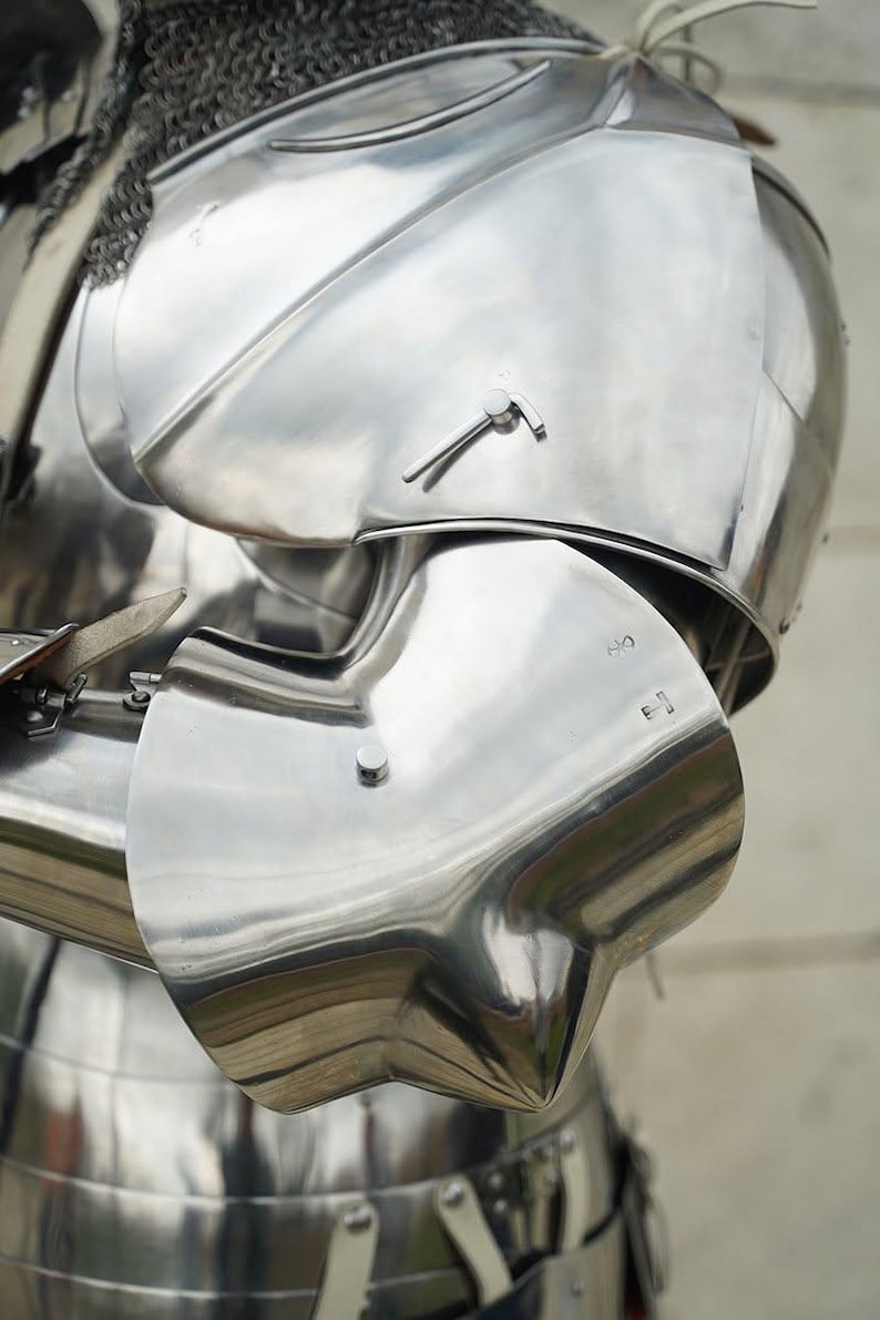 May include: A polished steel suit of armour featuring a rounded shoulder pauldron and a curved elbow couter. The metallic surface reflects light, showing smooth contours, rivets, and a chainmail collar visible at the top of the shoulder piece.