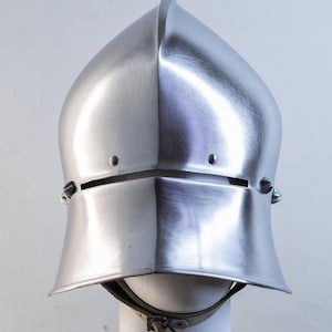 Könnte beinhalten: Ein polierter, silberfarbener Sallet-Helm aus Stahl mit spitzer Krone, einem horizontalen Sehschlitz und einem ausgestellten Nackenschutz. Der Helm verfügt über zwei kleine Nieten an der Vorderseite und einen Kinnriemen aus Leder an der Basis.