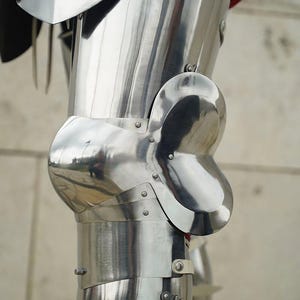 May include: A polished steel leg armour set featuring a curved thigh plate, a prominent rounded knee cop with side wings, and a lower greave section. The metal components are connected with visible rivets and straps, showing a reflective surface.