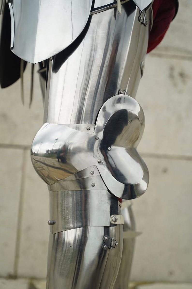 May include: A polished steel leg armour set featuring a curved thigh plate, a prominent rounded knee cop with side wings, and a lower greave section. The metal components are connected with visible rivets and straps, showing a reflective surface.