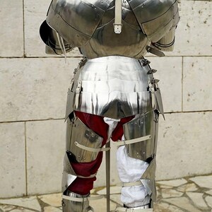 May include: A full suit of polished steel plate armour viewed from the back, featuring a rounded helmet, articulated shoulder pauldrons, a segmented breastplate, and greaves. The armour is worn over red and white fabric leg coverings.