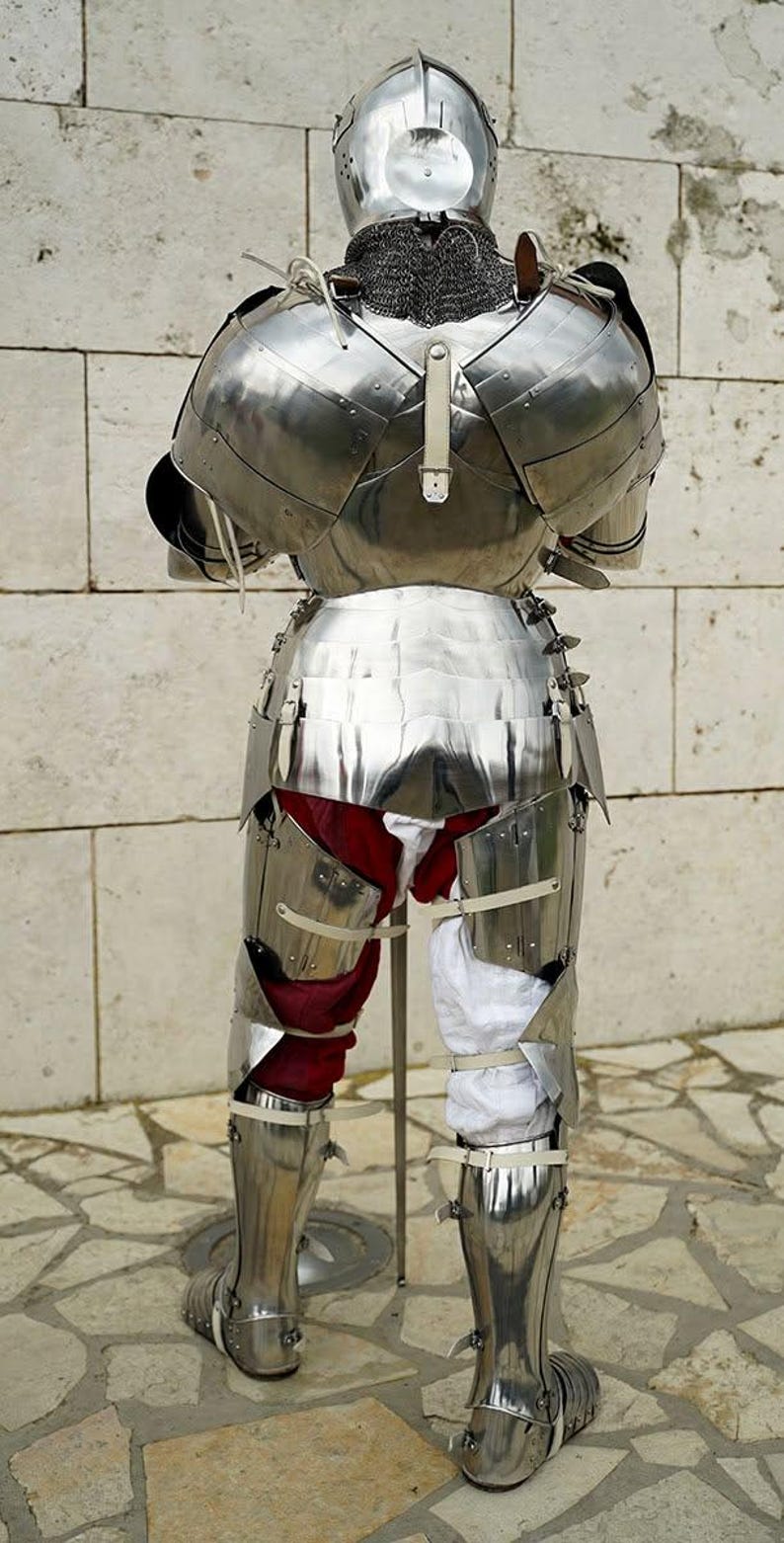 May include: A full suit of polished steel plate armour viewed from the back, featuring a rounded helmet, articulated shoulder pauldrons, a segmented breastplate, and greaves. The armour is worn over red and white fabric leg coverings.
