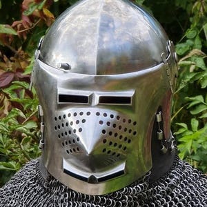 May include: A polished steel bascinet helmet featuring a pointed crown, a hinged visor with rectangular eye slits, and a perforated face plate. The helmet is worn over a silver-toned chainmail aventail that covers the neck and shoulders.