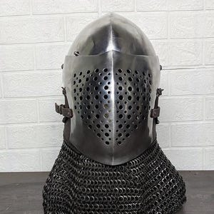 Puede incluir: Un casco de tipo bacinete de acero pulido con una corona puntiaguda y una visera abatible con numerosos orificios de ventilación circulares. El casco cuenta con una correa de cuero marrón oscuro y una camal de malla metálica oscura para el cuello.
