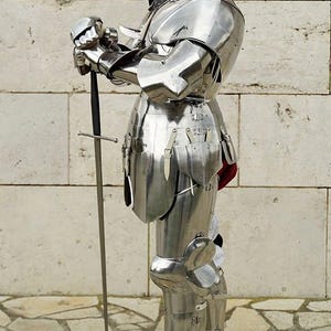May include: A full suit of polished silver-coloured steel plate armour featuring a closed helmet with a visor, articulated shoulder, arm, and leg pieces, and a metal gauntlet holding a long, slender sword with a straight crossguard and black hilt.