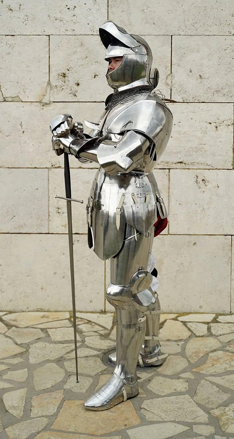 May include: A full suit of polished silver-coloured steel plate armour featuring a closed helmet with a visor, articulated shoulder, arm, and leg pieces, and a metal gauntlet holding a long, slender sword with a straight crossguard and black hilt.