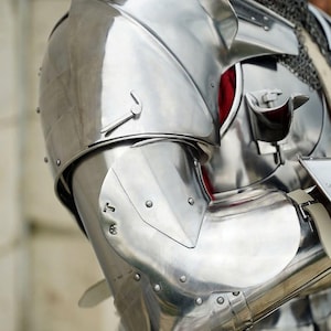 May include: A polished steel suit of armour featuring a rounded shoulder pauldron, a segmented arm guard, and a metal elbow cop. The armour includes a chainmail neck covering and a chest plate with a visible red fabric lining underneath the metal.