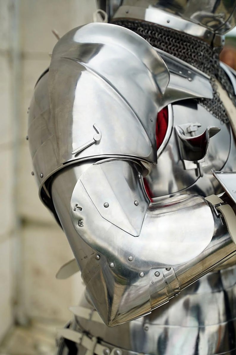May include: A polished steel suit of armour featuring a rounded shoulder pauldron, a segmented arm guard, and a metal elbow cop. The armour includes a chainmail neck covering and a chest plate with a visible red fabric lining underneath the metal.