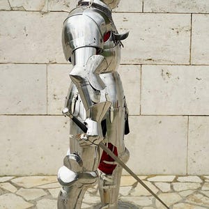 May include: A full suit of polished silver-coloured metal plate armour featuring a helmet with a visor, articulated shoulder, arm, and leg pieces, and a metal gauntlet holding a long, slender sword with a dark handle and a metallic crossguard.