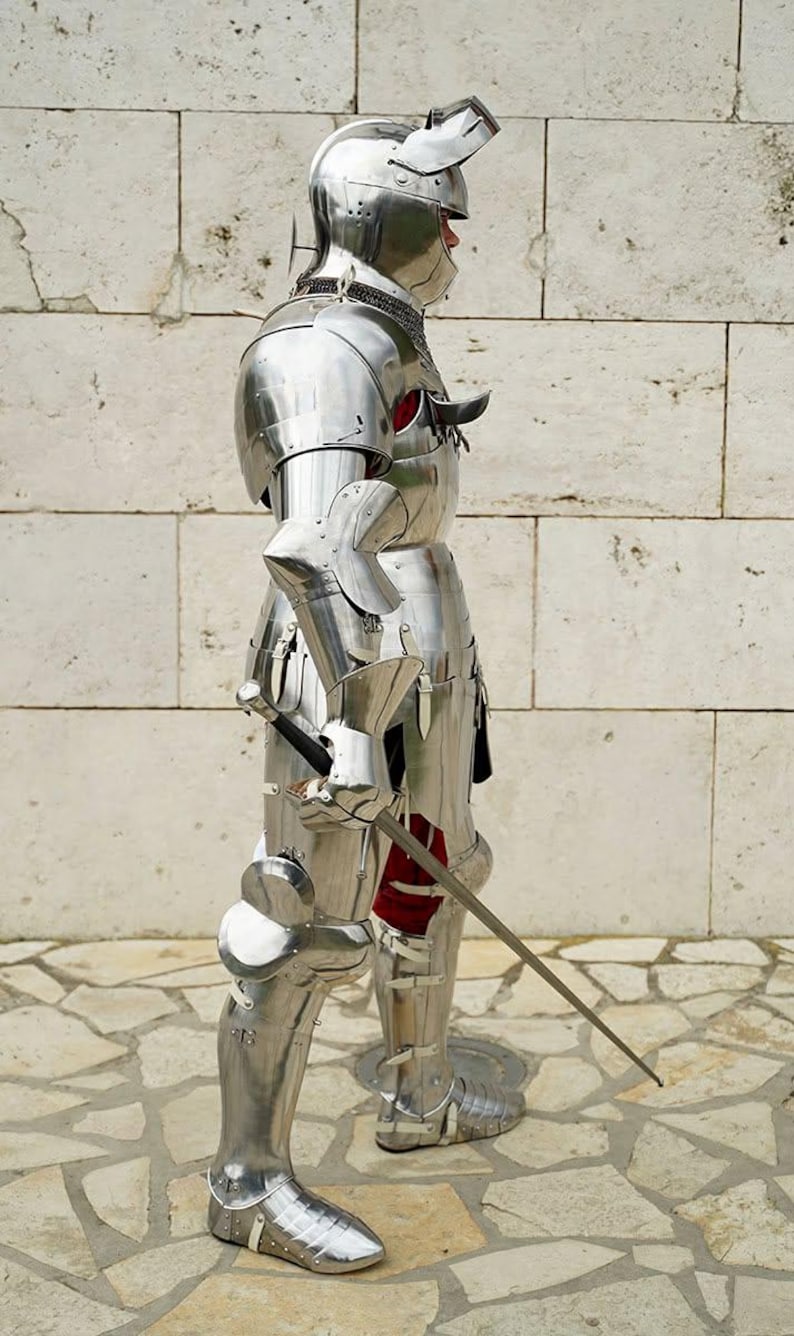 May include: A full suit of polished silver-coloured metal plate armour featuring a helmet with a visor, articulated shoulder, arm, and leg pieces, and a metal gauntlet holding a long, slender sword with a dark handle and a metallic crossguard.