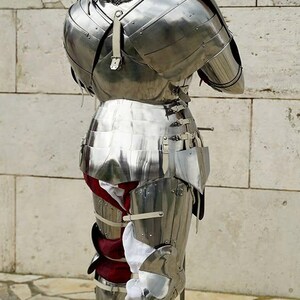May include: A full suit of polished steel plate armour viewed from the back, featuring a rounded helmet, articulated shoulder pauldrons, a segmented breastplate, and leg greaves. White and red fabric padding is visible beneath the metal plates.