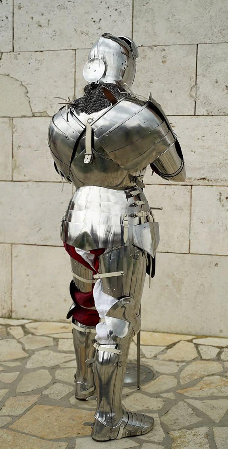 May include: A full suit of polished steel plate armour viewed from the back, featuring a rounded helmet, articulated shoulder pauldrons, a segmented breastplate, and leg greaves. White and red fabric padding is visible beneath the metal plates.