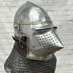 Op de afbeelding: Een middeleeuwse helm van gepolijst staal met een puntig vizier met ventilatiegaten, geaccentueerd door getextureerde koperen randen. De helm is voorzien van een bruine leren nekband en een beschermende maliënkolder aan de onderzijde.