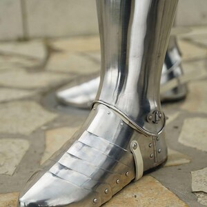 May include: A polished steel greave and sabaton set covering the lower leg and foot. The armour features articulated metal plates over the foot, secured with small rivets and a light-coloured leather strap at the ankle, reflecting a bright metallic sheen.