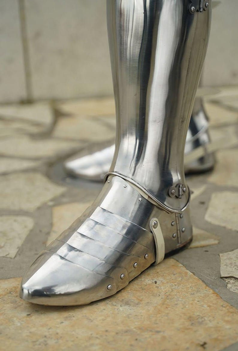 May include: A polished steel greave and sabaton set covering the lower leg and foot. The armour features articulated metal plates over the foot, secured with small rivets and a light-coloured leather strap at the ankle, reflecting a bright metallic sheen.