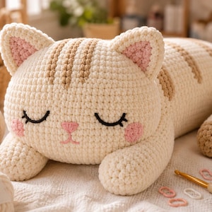 May include: A cream-coloured crocheted cat plush toy with light brown stripes on its head and back. The cat has closed eyes, a pink stitched nose and mouth, and pink circular cheeks. It is lying down with its front paws extended forward.