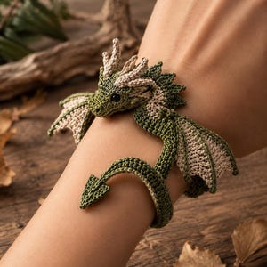 Op de afbeelding: Een gehaakte drakenarmband gemaakt van groen en beige garen. Het ontwerp bevat een gedetailleerde drakenkop met kleine hoorns, getextureerde vleugels en een lange, gebogen staart die om de pols krult om een cirkelvormige band te vormen.