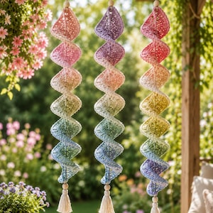 May include: Three hanging spiral decorations made of crocheted yarn in a gradient of colors including pink, purple, yellow, green, and blue. Each piece features a wooden bead at the top and a cream-colored tassel at the bottom, suspended by string.