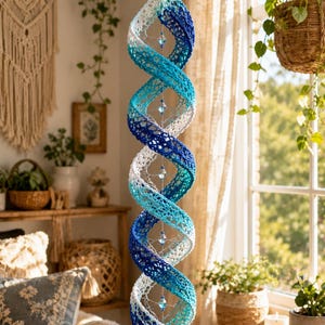 May include: A hanging spiral ornament featuring a crochet design with a gradient of white, light blue, and dark blue yarn. The structure is held together by a central vertical string adorned with small, clear, faceted glass beads.