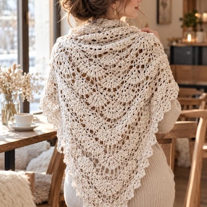 May include: A cream-colored triangular shawl featuring an intricate openwork crochet pattern with repeating fan-shaped motifs and a scalloped edge. The lightweight textile is draped over the shoulders, showcasing a detailed, airy, and textured design.