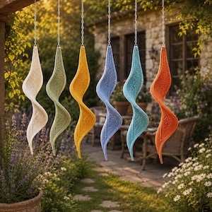 May include: Six spiral-shaped hanging decorations made of textured, crocheted fabric in cream, sage green, mustard yellow, periwinkle blue, turquoise, and burnt orange. Each piece is suspended from a thin silver-colored metal chain.