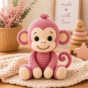 May include: A crocheted monkey toy in shades of dusty pink and cream. The monkey features a round head, large circular ears with cream centres, black plastic eyes, a stitched smile, and a curled tail. It is crafted with a textured yarn finish.