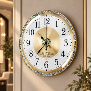 Könnte beinhalten: Eine runde Wanduhr mit goldenem Rahmen und einem Rand aus irisierenden Kristallen. Das Zifferblatt hat einen weißen Hintergrund mit schwarzen Ziffern, eine goldene Mitte und schwarze Zeiger. Der Text lautet The Wilsons EST. 2026.