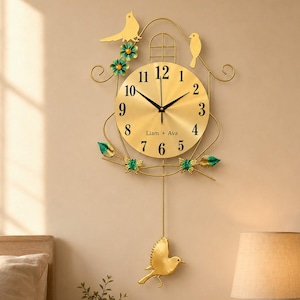Könnte beinhalten: Eine goldfarbene Wanduhr mit rundem Zifferblatt, schwarzen Ziffern und der Aufschrift Liam + Ava. Die Uhr ist mit metallenen Vogel- und Blumenmotiven in Gold und Türkis verziert und besitzt ein Pendel in Form eines kleinen Vogels.