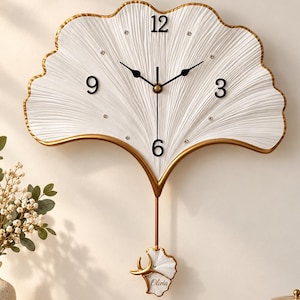 May include: A white ginkgo leaf-shaped wall clock with a gold-toned border, black hands, and black numerals at 3, 6, 9, and 12. The clock features a pendulum with a smaller leaf accent bearing the name Olivia in cursive script.