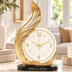 Puede incluir: Un reloj de mesa con esfera de mármol blanco, números y manecillas doradas, enmarcado por una estructura dorada con forma de pluma. El reloj se asienta sobre una base rectangular negra con el texto EMMA & NOAH impreso en color oro.