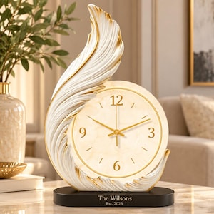 Puede incluir: Un reloj de mesa con un marco en forma de pluma blanca y dorada que rodea una esfera circular con manecillas doradas y los números 3, 6, 9 y 12. La base negra tiene grabada la inscripción "The Wilsons" y "Est. 2026" en blanco.
