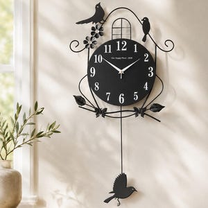 May include: A black wall clock with a circular face, white numerals, and white hands. The clock features a metal frame with silhouettes of birds, flowers, and butterflies. A pendulum with a bird silhouette hangs below. Text reads: Our Happy Place - 2026.