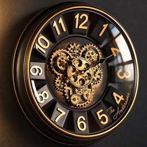 May include: A round wall clock with a black frame and gold-coloured numerals from 1 to 12. The centre features exposed, rotating gold-toned gears against a dark background. The text The Andersons is printed in gold script near the number 4.