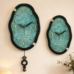 May include: Two irregular oval wall clocks with black frames and textured teal and gold faces. Each clock features gold numerals 3, 6, 9, and 12, black hands, and the text Olivia & James 06.14.2026. The larger clock includes a black geometric pendulum.