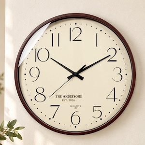 May include: A round wall clock with a dark brown frame and a cream-colored face. It features black Arabic numerals, black hour and minute hands, and a thin second hand. The text reads THE ANDEEFSOHS EST. 2026 QUARTZ in the center of the clock face.