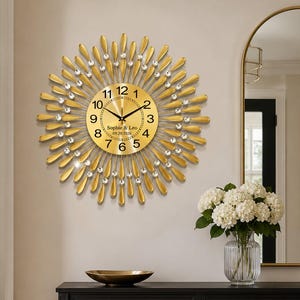 Könnte beinhalten: Eine runde goldene Wanduhr mit einem Strahlenkranz-Design aus strahlenförmigen Metallblütenblättern, die mit klaren Kristallen verziert sind. Das goldene Zifferblatt zeigt schwarze arabische Ziffern, schwarze Zeiger und den Text Sophie & Leo 09.20.2026.