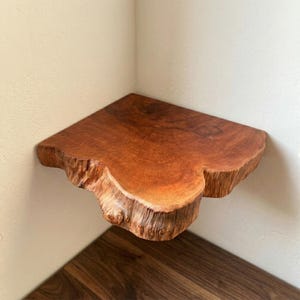 May include: A corner shelf made from a thick, solid slab of natural wood with a warm reddish-brown tone. The shelf features a prominent, irregular live edge that showcases the raw, textured bark and organic grain patterns of the wood.