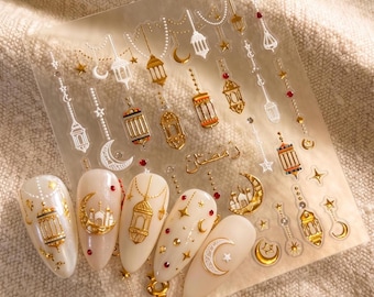 May include: A sheet of adhesive nail decals featuring gold and white designs of lanterns, crescent moons, stars, and mosque silhouettes. The sheet includes individual nail-shaped stickers with intricate metallic patterns and Arabic calligraphy.