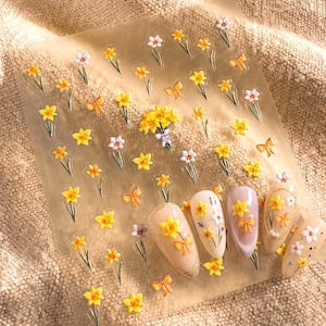 May include: A clear sheet of adhesive nail decals featuring various yellow and white flower designs, small orange bows, and a central bouquet. Four artificial nail tips are displayed at the bottom, decorated with matching floral and bow stickers.