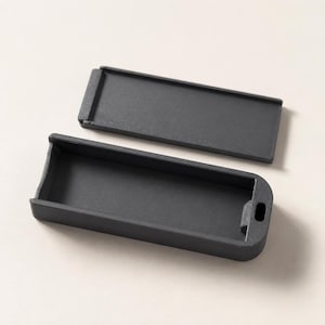 May include: A rectangular matte black storage container with a matching sliding lid. The base features a hollow interior with a small circular opening on one end, while the lid is a flat, solid piece designed to fit securely over the container.