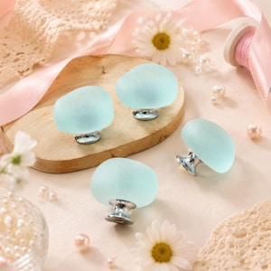 May include: Four cabinet knobs featuring smooth, frosted light blue glass tops with a rounded, pebble-like shape. Each knob is attached to a polished silver-toned metal base, designed for mounting onto furniture drawers or cabinet doors.